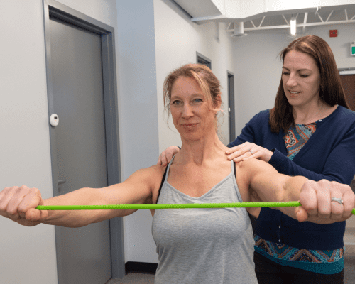 She is lying on a massage table with a green cord stretched across her body, while another woman is standing behind her, providing the massage