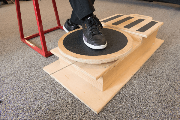 a person wearing black shoes standing on a platform, which appears to be a balance board or a small wooden stool